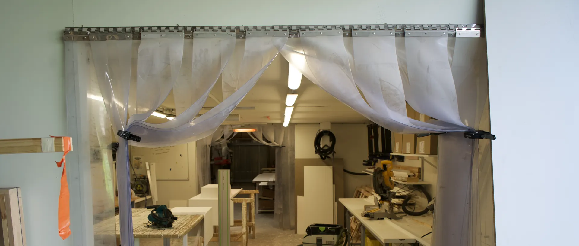 Atelier de montage vu à travers son rideau à lanières : établis, scie à onglet et meubles en cours d'assemblage